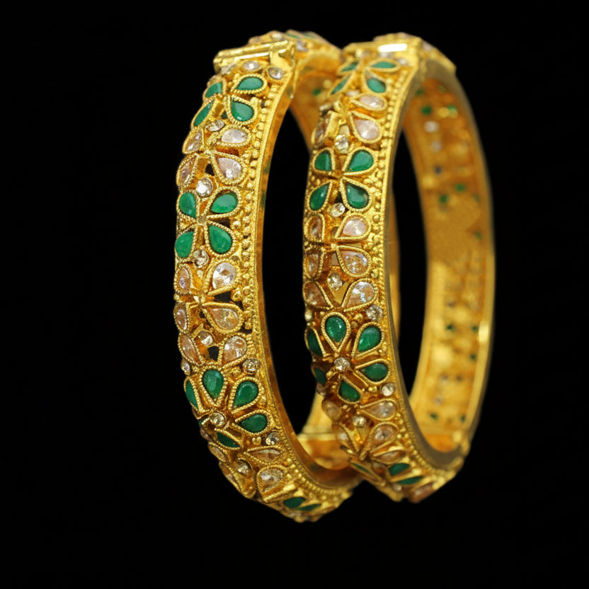 Gold bangles with green stones on a dark background, featuring the brand 'Saheli'.