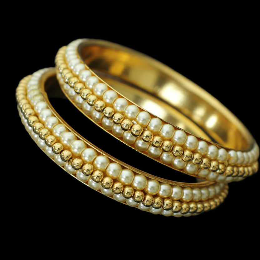 Two gold and pearl bangles on a reflective surface with 'Saheli' branding.