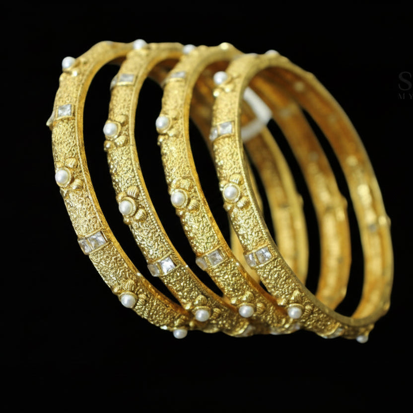 Gold bangles with pearl embellishments on a dark background, featuring the brand 'SAHELI'.