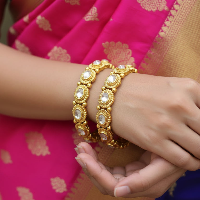 Gold bangles with intricate designs on a gray background, featuring the brand 'Saheli'.