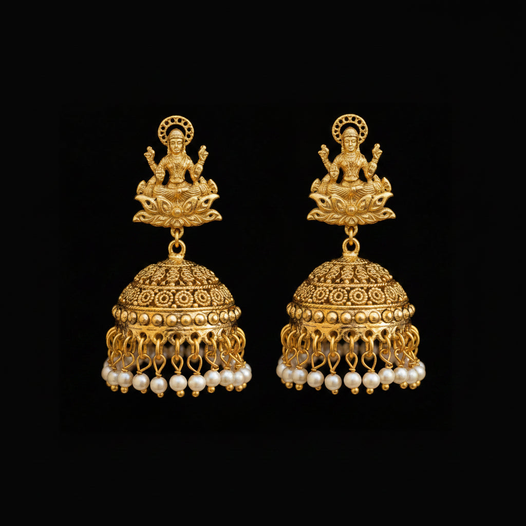 A pair of gold-toned Indian jhumka earrings with detailed filigree work, small white pearl bead drops, and deity motifs at the top, displayed on a black Y-shaped stand against a gray background. The brand name 'SAHELI MY BEST FRIEND' is visible at the top center.