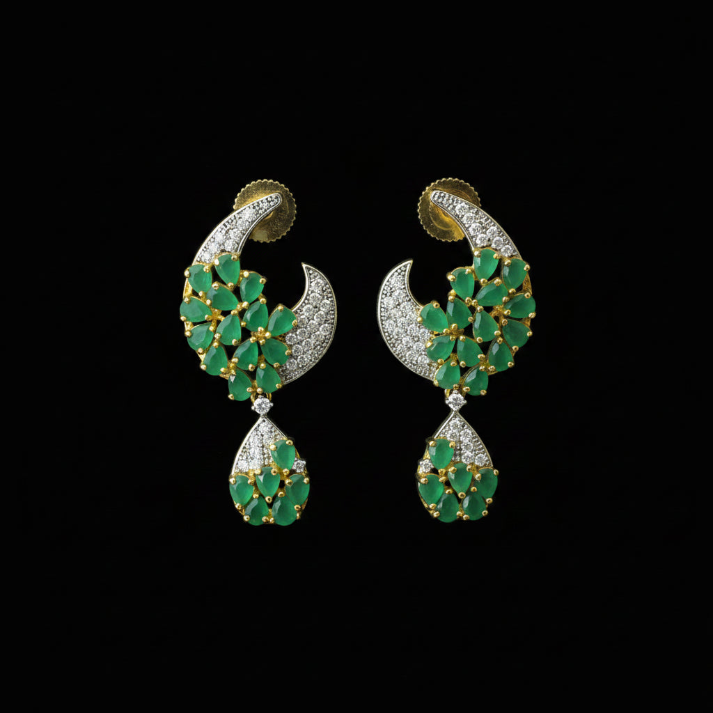 A pair of gold-plated crescent-shaped earrings with green stone clusters and small clear stones, displayed on a dark reflective surface. The SAHeli logo is visible in the top left corner.