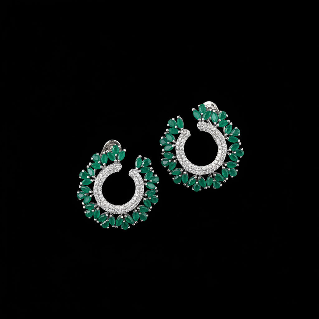 A pair of crescent-shaped hoop earrings with green emerald-cut stones and a silver-toned center set with clear stones, displayed on a dark reflective surface with the SAHELI brand logo above.