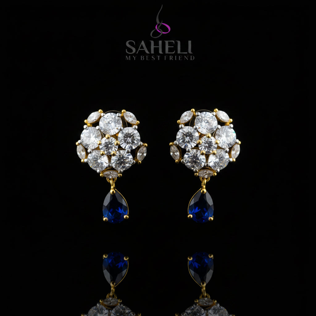A pair of gold-plated stud earrings with clear round-cut stones in a floral cluster design, each hanging from a teardrop-shaped dark blue stone, displayed on a black jewelry stand against a gray background with the Saheli brand logo at the top.
