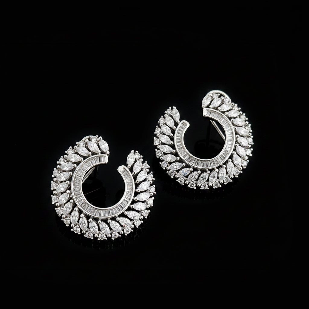 A pair of silver crescent-shaped hoop earrings adorned with clear pavé stones, displayed on a dark reflective surface with the SAHELI MY BEST FRIEND logo in the upper center.