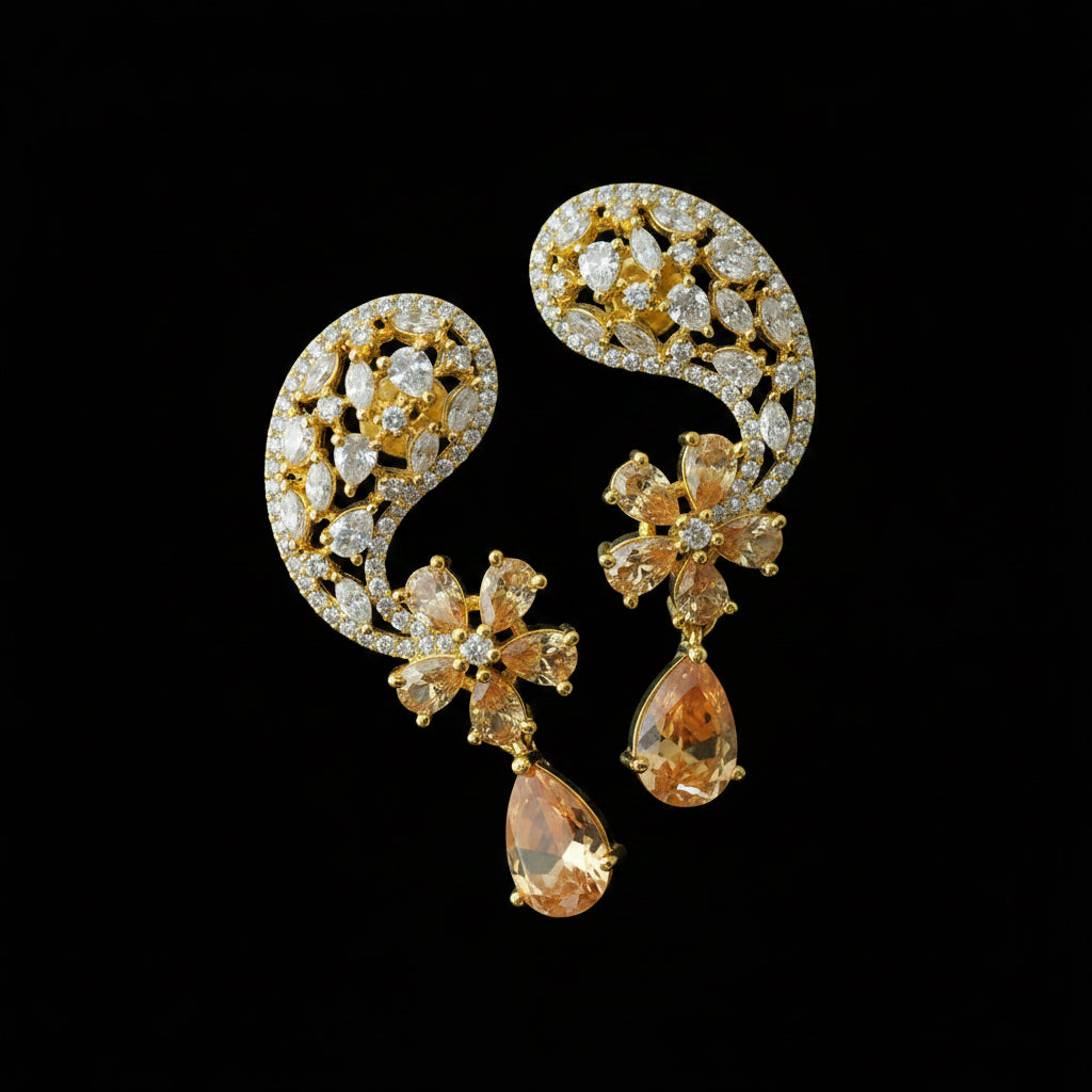 A pair of ornate gold-plated dangle earrings embellished with clear crystals and peach-colored teardrop rhinestones, displayed on a dark reflective surface with the Saheli brand logo in the top left corner.