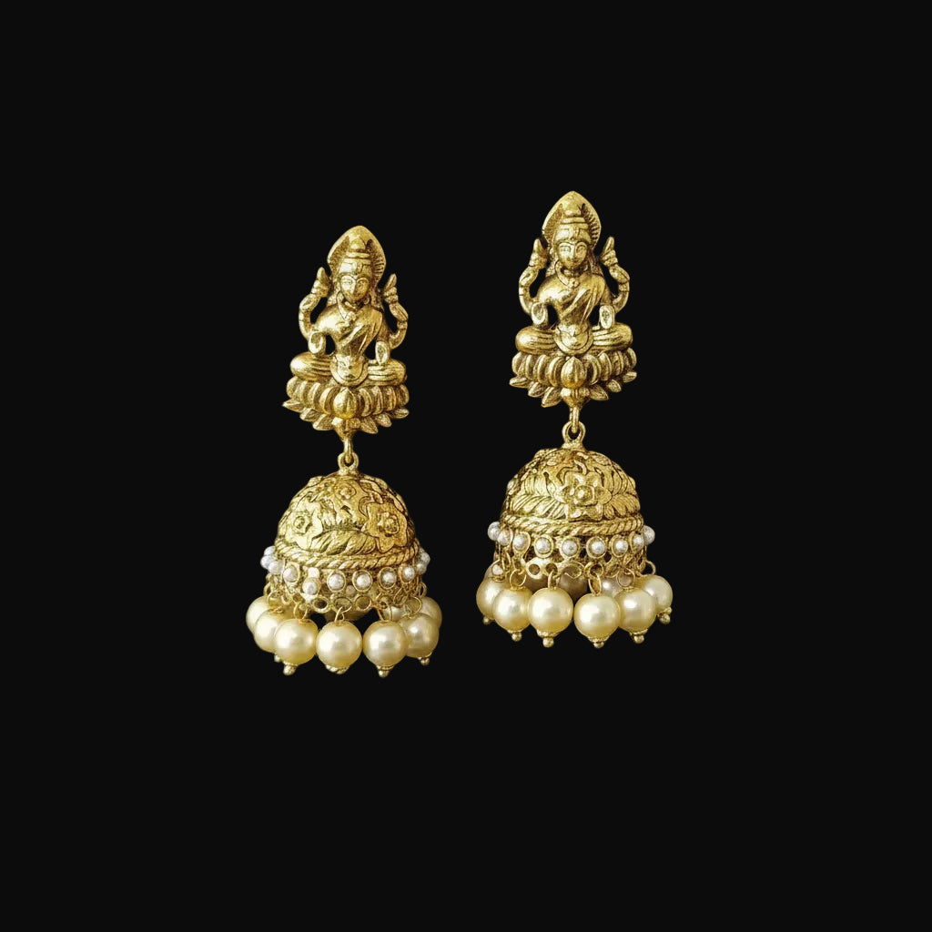 A pair of gold-toned traditional Indian Jhumka earrings displayed on a black jewelry stand. Each earring features a deity figurine at the top, an ornate filigree dangle, and a row of small white faux pearls at the bottom.