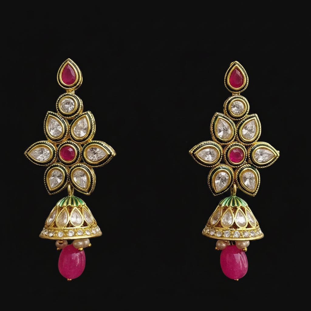 A pair of gold-plated Indian jhumka earrings with pink stone accents, clear crystals, and green enamel details, displayed on a black Y-shaped stand against a gray background. The brand logo 'SAHELI MY BEST FRIEND' is visible at the top.