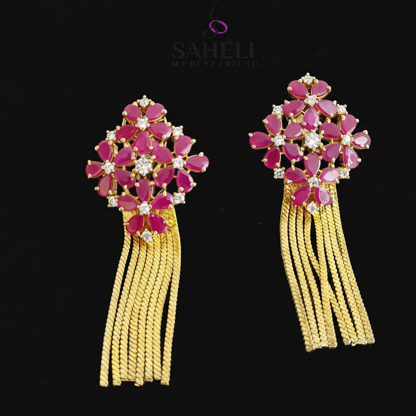 Pair of earrings with pink gemstones and yellow tassels on a dark background, branded 'Saheli'.