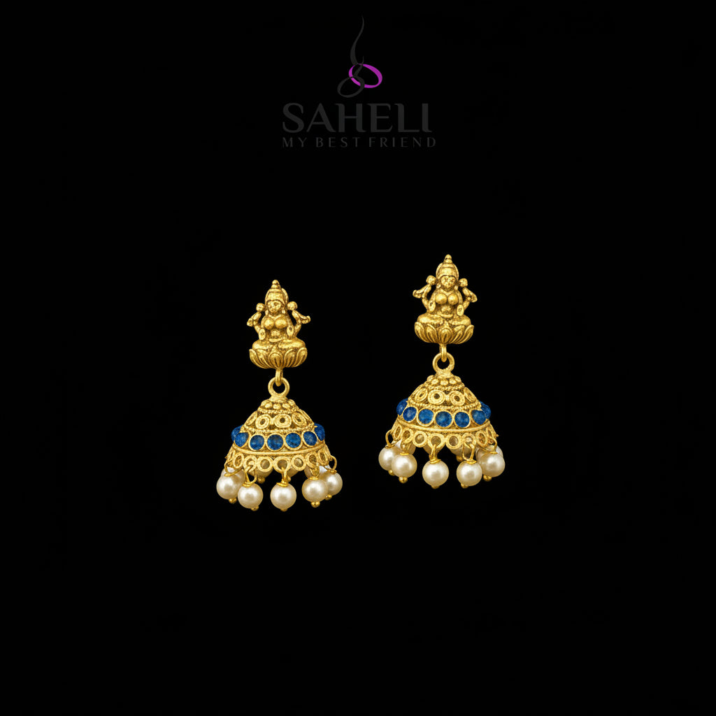 A pair of gold-toned traditional Indian jhumka earrings with blue stone accents and white pearl drops, displayed on a gray Y-shaped stand. The brand logo 'SAHELI MY BEST FRIEND' is visible at the top center.