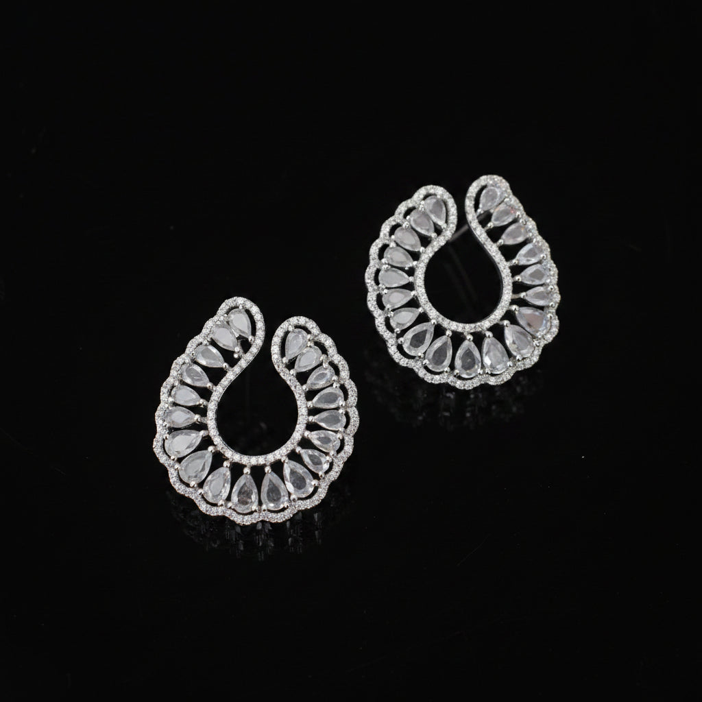 A pair of silver-toned teardrop-shaped earrings with intricate filigree cutouts and clear stones, displayed on a dark reflective surface.