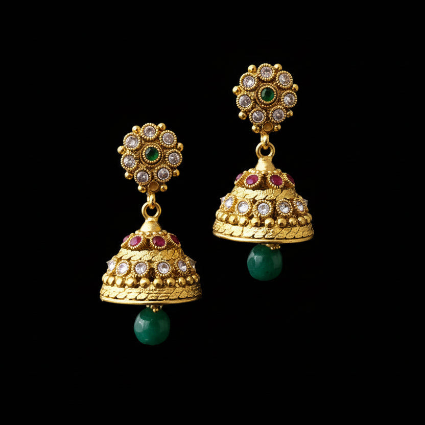 Gold jhumka earrings with green and red stones on a gray stand, branded 'Saheili'.