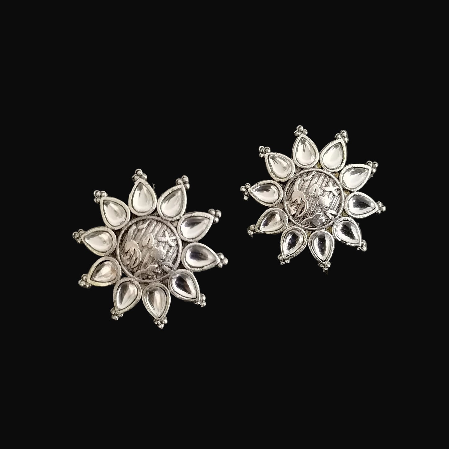 Silver floral earrings on a dark background with 'SAHELI' branding.