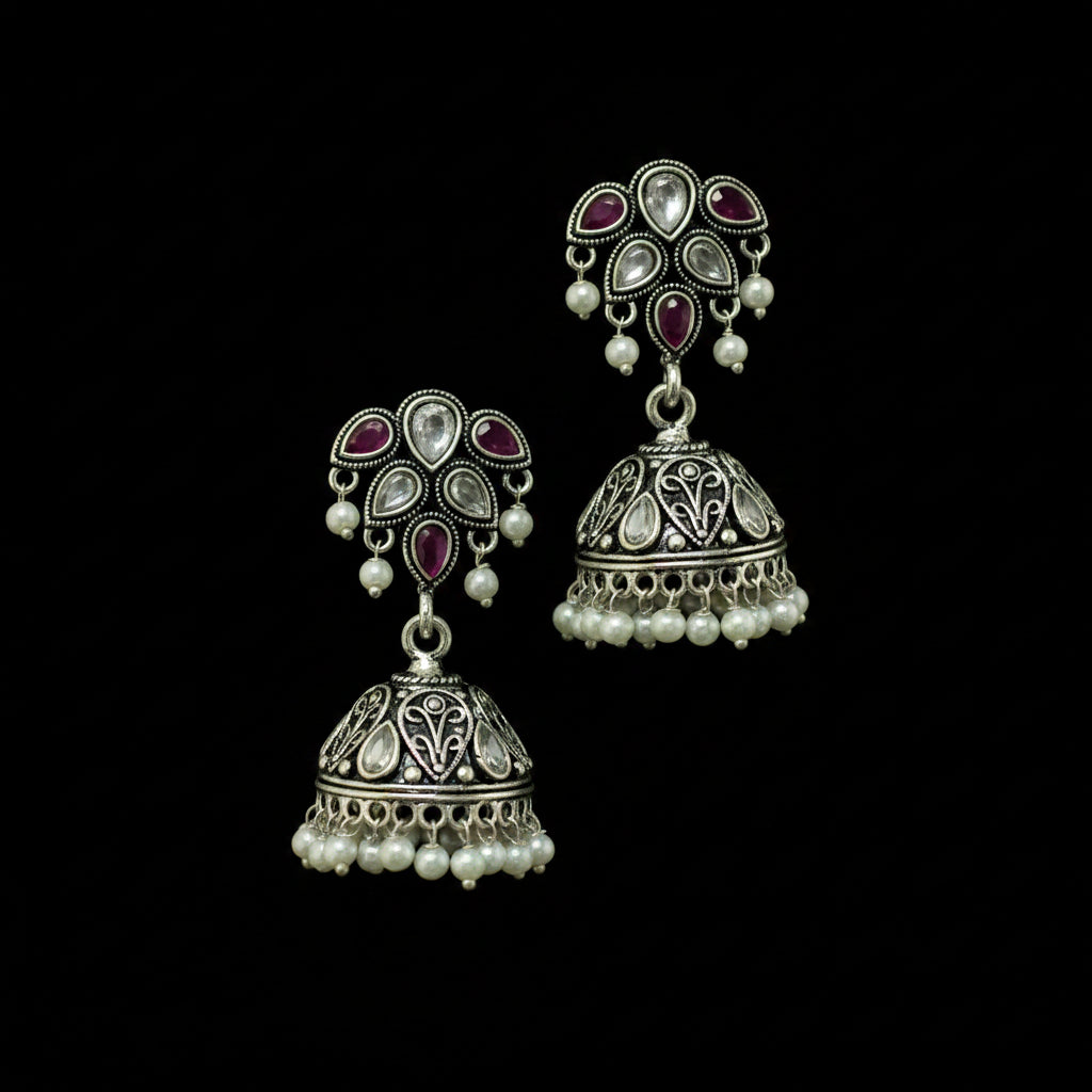 A pair of traditional Indian silver jhumka earrings with pink stone accents and white pearl bead fringe, displayed on a black Y-shaped stand against a gray background.