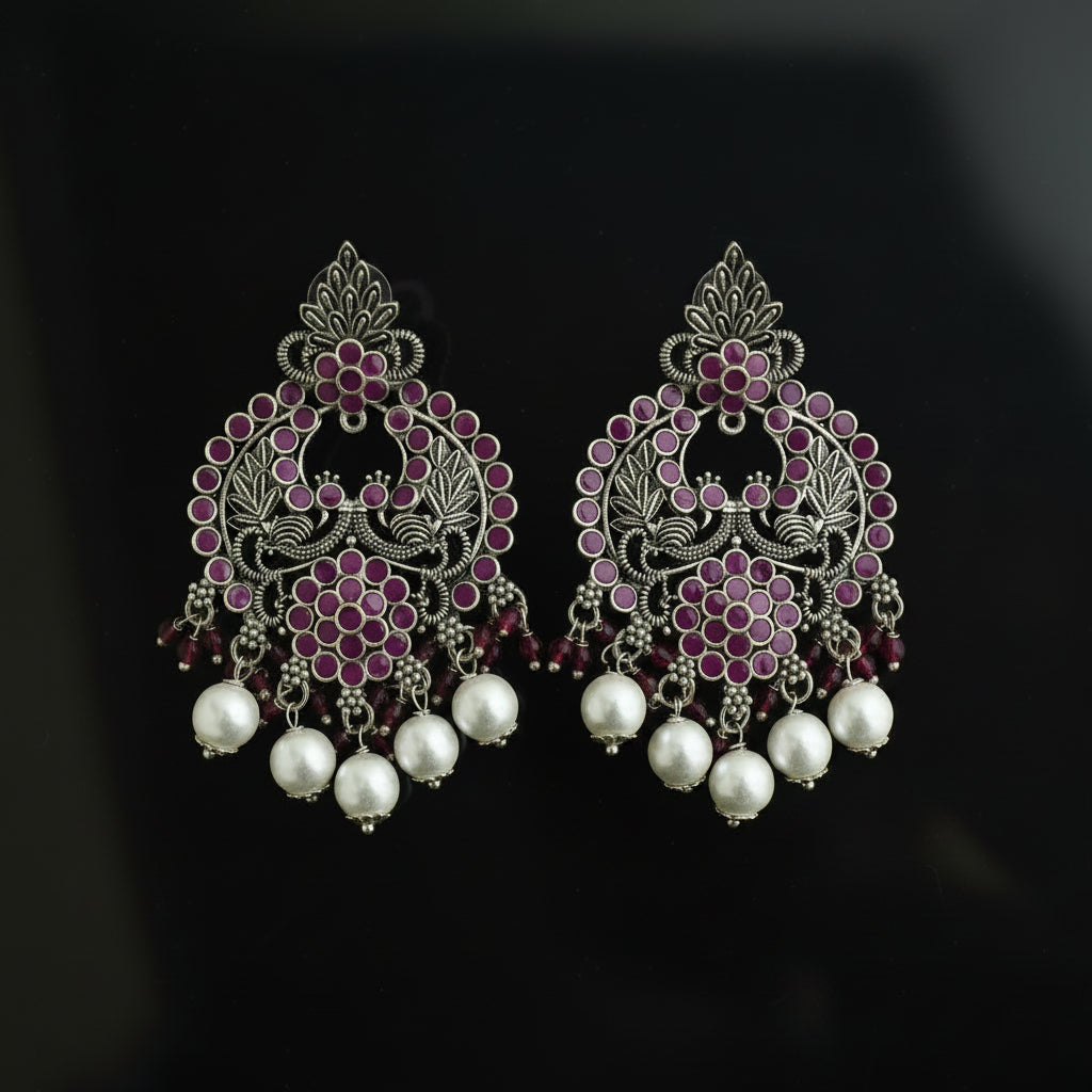 A pair of ornate silver-toned filigree earrings decorated with pink and purple stones and dangling white pearl beads, displayed on a dark reflective surface.