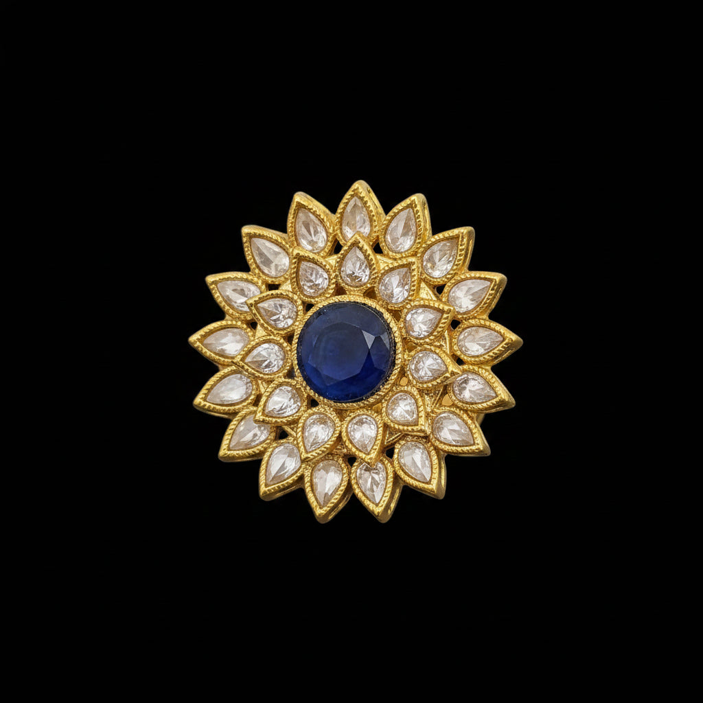 A gold-toned floral brooch with a central deep blue oval stone and surrounding clear teardrop-shaped stones, displayed on a reflective dark surface with the SAHeli logo in the top left corner.
