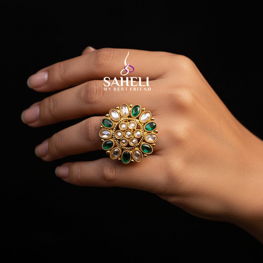 A large gold-toned statement ring with green and clear stones, displayed on a black velvet hand against a black background. The brand logo 'SAHELI MY BEST FRIEND' is visible on the ring.