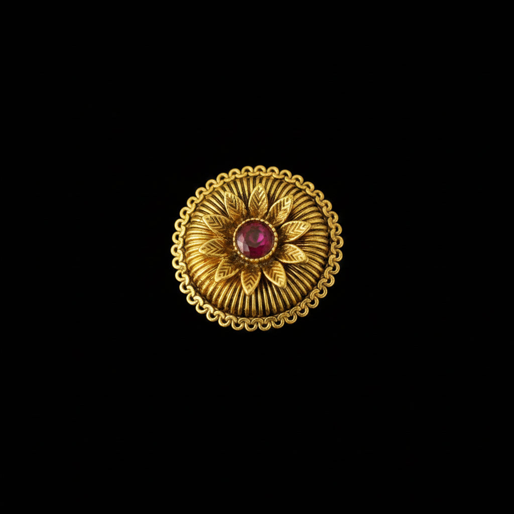 A gold ring with a circular sunburst design and a central red gemstone, displayed on a black gloved hand against a dark background.