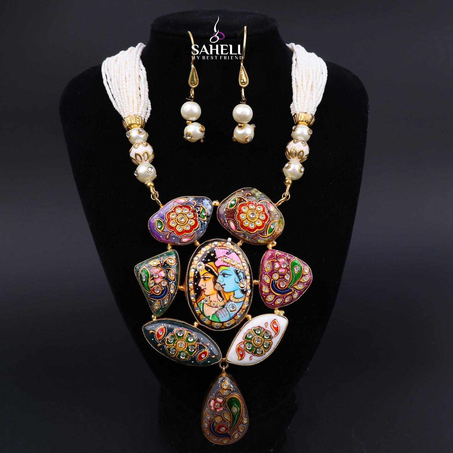 A traditional Indian statement necklace and matching drop earrings displayed on a black velvet jewelry bust. The necklace features a central oval pendant depicting Lord Krishna, surrounded by colorful floral and paisley enamel-style panels in red, green, blue, and gold, accented with white tassel strands and pearl-like beads.