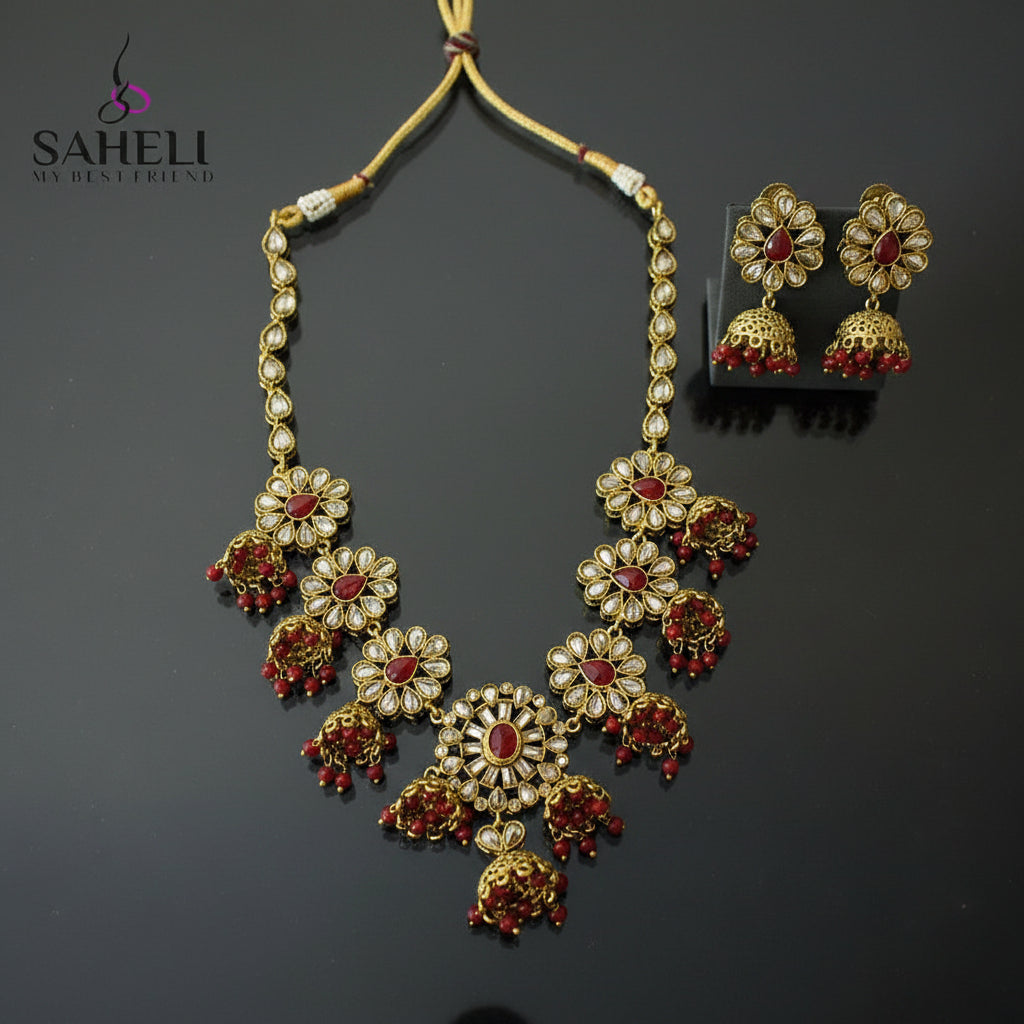 Gold-toned traditional Indian jewelry set featuring a statement necklace and matching earrings with red stones and white stones, displayed on a dark reflective surface. The SAHELI MY BESTFRIEND logo is visible in the top left corner.