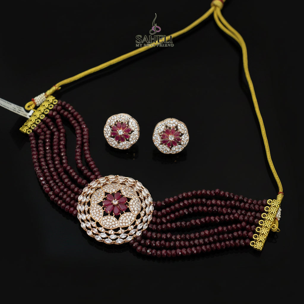 Jewelry set with maroon beads and gold accents on a dark surface, featuring the Saheli brand.