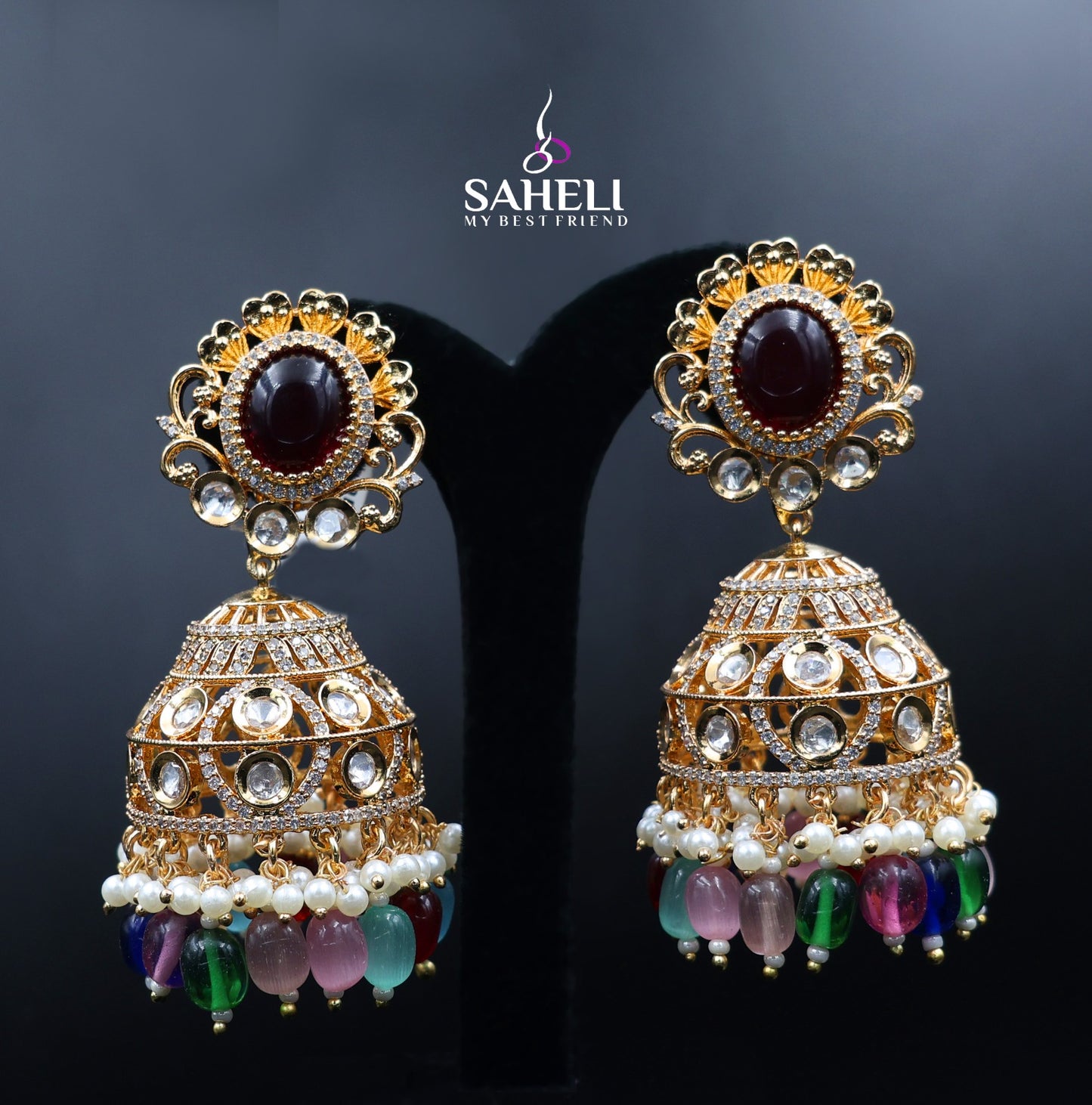 Ornate gold earrings with gemstones and pearls on a dark background, featuring the brand 'Saheli'.