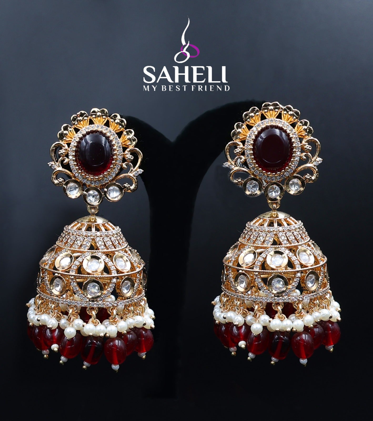 Ornate earrings with red stones and gold accents on a dark background, featuring the brand 'Saheli'.