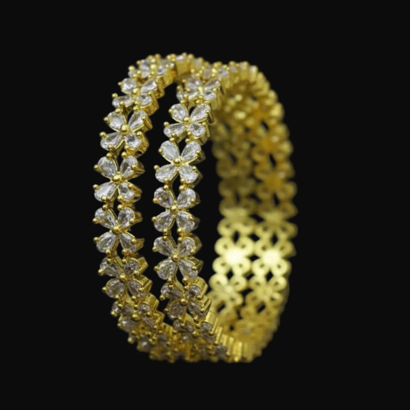 Gold bangles with clear stones on a black background, featuring the brand 'Saheli'.