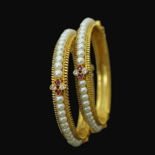 Gold and pearl bangles with red stones on a black background, featuring the brand 'Saheli'.