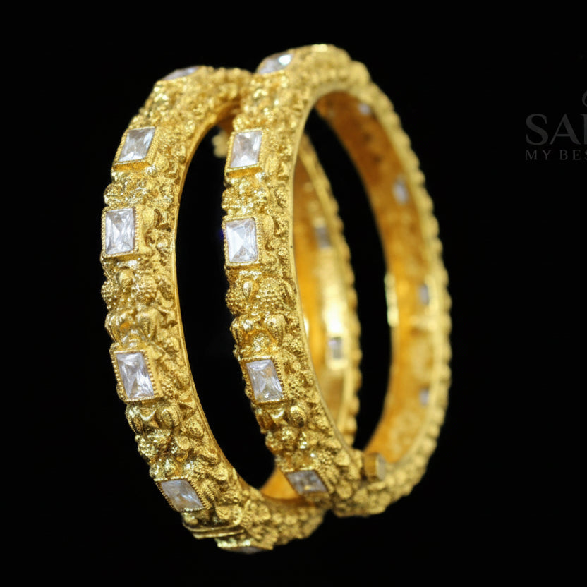 Gold bangles with embedded stones on a dark background, featuring the brand 'Saheli'.