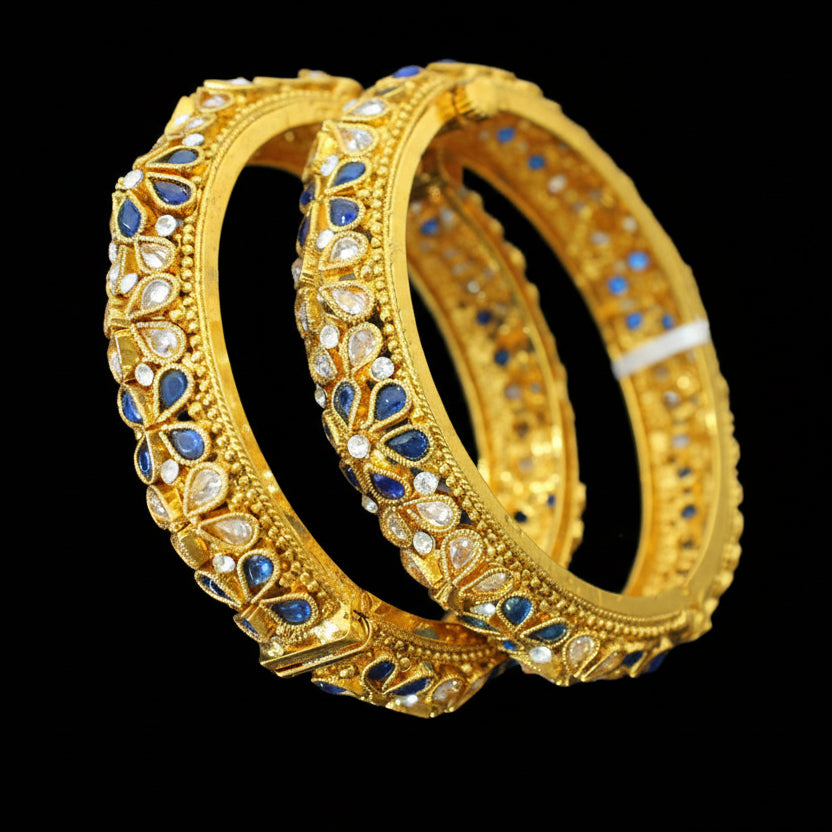 Gold bangles with blue and white stones on a gray surface, branded 'SAHELI'.