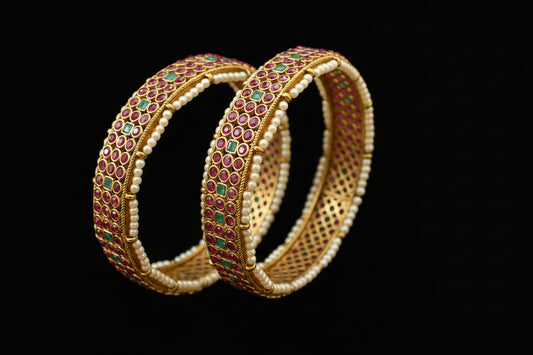 Gold bangles with red and green stones on a black stand, branded 'Saheli'.