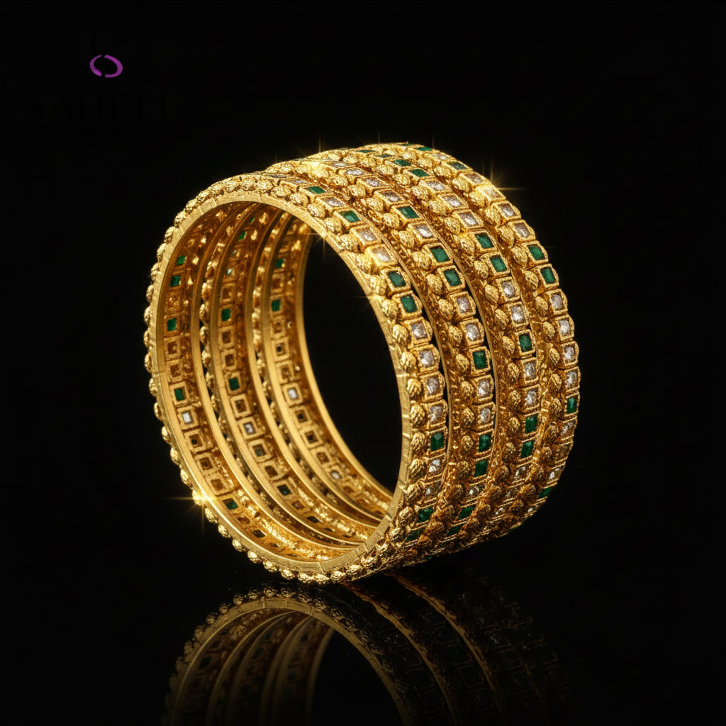 Gold bangles with green stones on a gray stand, branded 'SAHELI'.