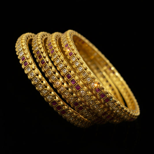 Set of gold bangles with red stones on a reflective surface, branded 'Saheli'.