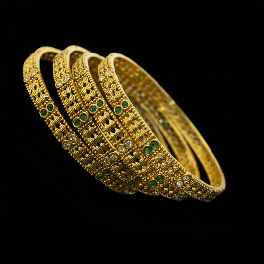 Gold bangles with green stones on a reflective surface, branded 'Saheli'.