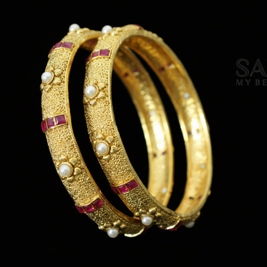 Gold bangles with pearls and red stones on a dark background, featuring the brand 'Saheili'.
