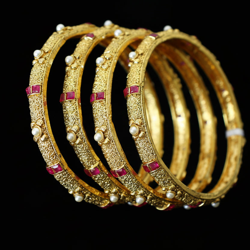 Gold bangles with red stones on a reflective surface, branded 'Saheli'.