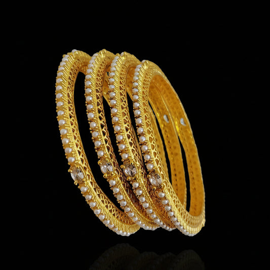 Set of gold bangles with pearl detailing on a dark surface, branded 'SAHELI'.