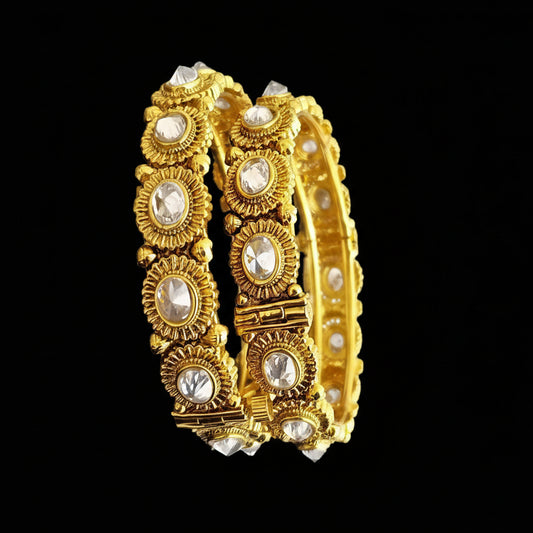 Gold bangles with intricate designs on a gray background, featuring the brand 'Saheli'.