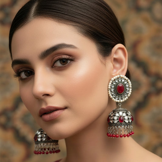 Pair of ornate earrings with red and silver details on a dark background, featuring the brand 'Saheli'.