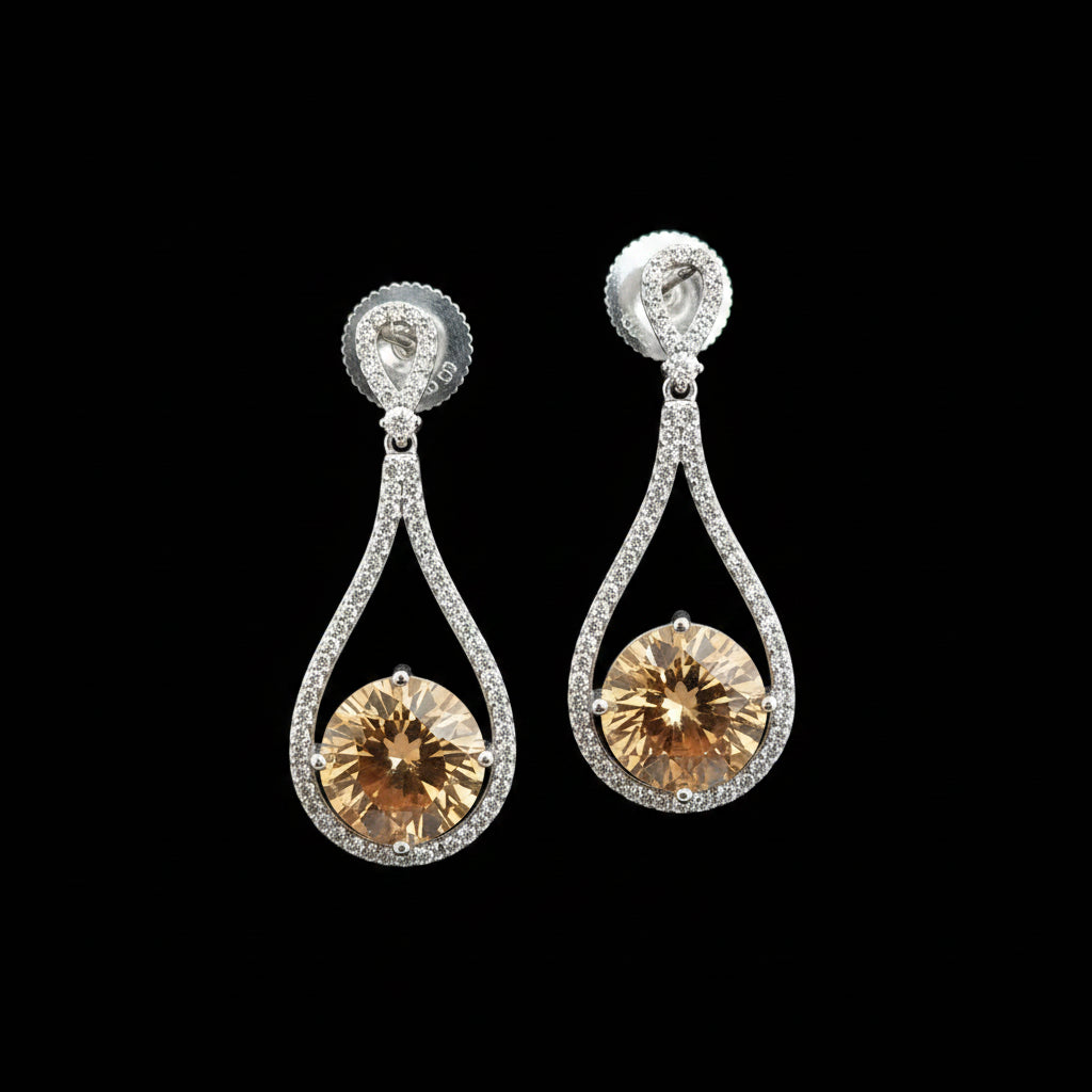 A pair of teardrop-shaped drop earrings with large round amber-colored center stones surrounded by small clear stones in silver-toned settings, displayed on a dark gray stand against a gray background. The brand name 'SAHELI MY BEST FRIEND' is visible in the top left corner.