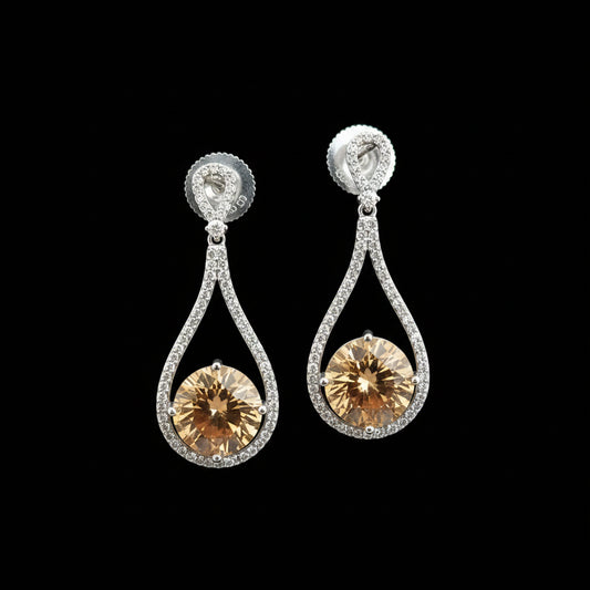 A pair of teardrop-shaped drop earrings with large round amber-colored center stones surrounded by small clear stones in silver-toned settings, displayed on a dark gray stand against a gray background. The brand name 'SAHELI MY BEST FRIEND' is visible in the top left corner.