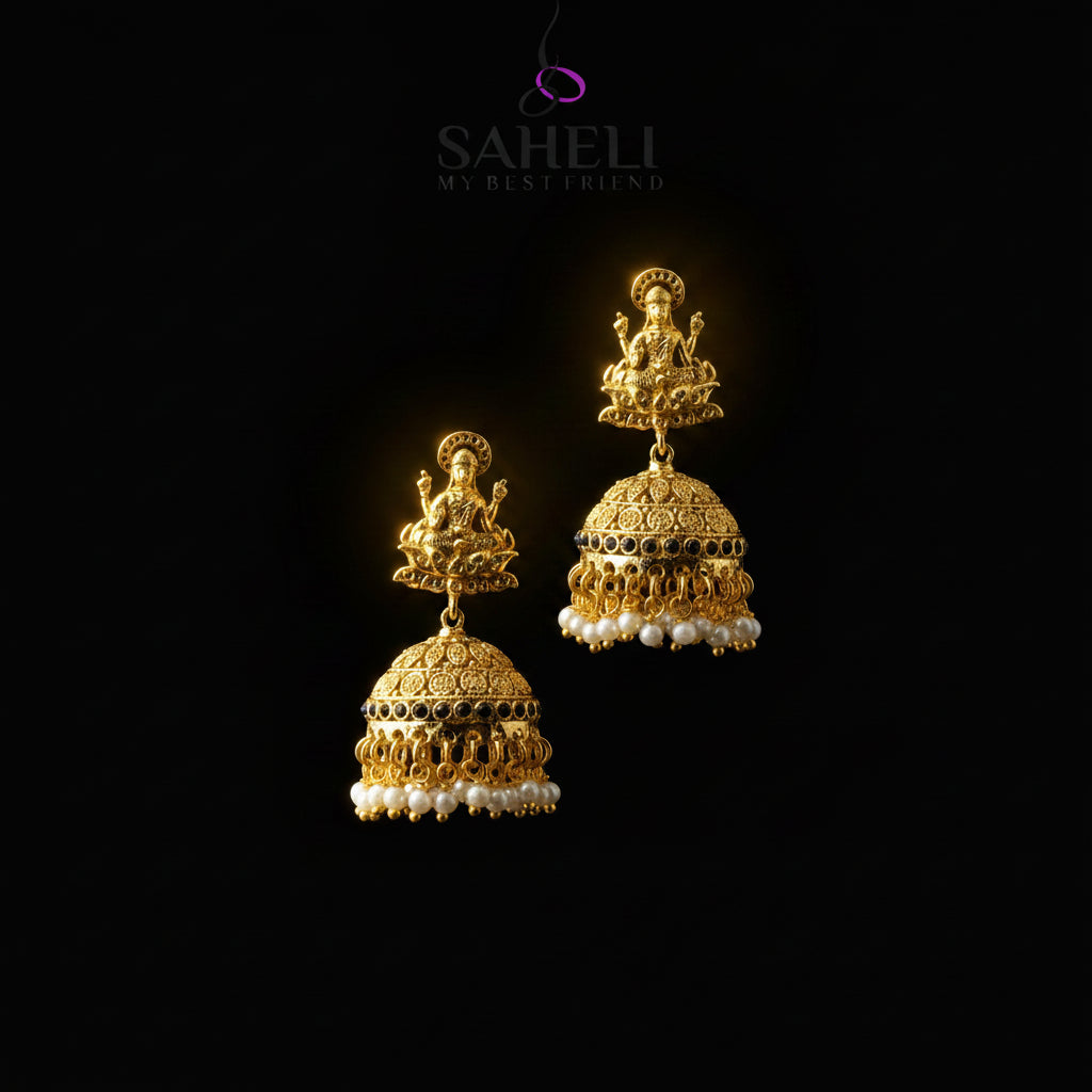 A pair of gold-toned traditional Indian jhumka earrings displayed on a black Y-shaped jewelry stand against a gray gradient background. The earrings feature detailed filigree work, small black bead accents, and rows of white pearl-like beads at the bottom.
