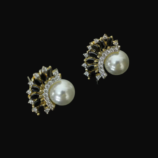 A pair of gold and black floral stud earrings with white pearls and clear crystals, displayed on a dark reflective surface with the SAHELI MY BEST FRIEND logo in the background.