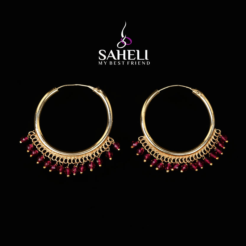Gold hoop earrings with red beads on a dark background, featuring the Saheli brand.