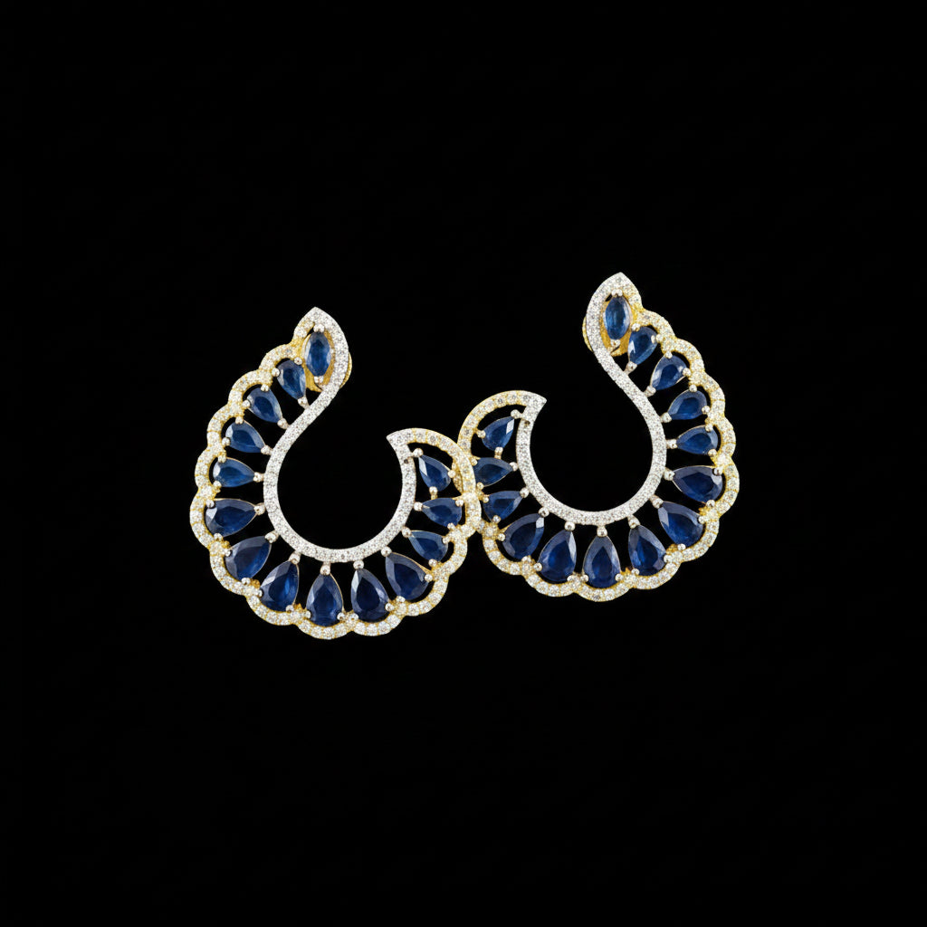 A pair of gold crescent-shaped earrings with deep blue teardrop stones and small clear accent stones, displayed on a dark reflective surface with the brand name 'SAHELI MY BEST FRIEND' above them.