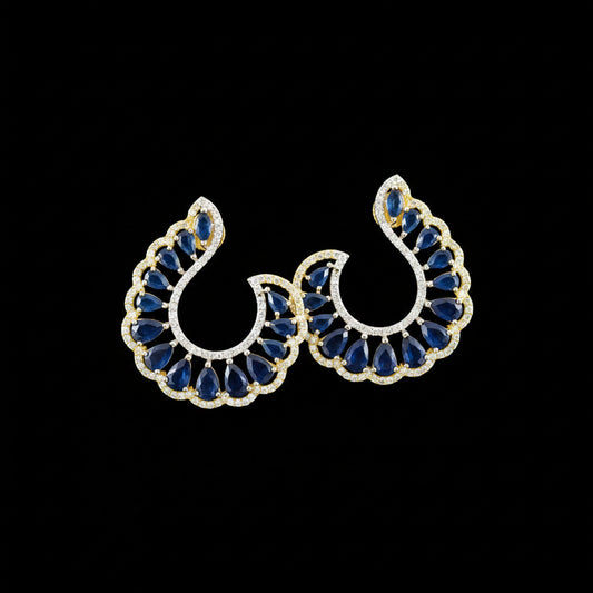 A pair of gold crescent-shaped earrings with deep blue teardrop stones and small clear accent stones, displayed on a dark reflective surface with the brand name 'SAHELI MY BEST FRIEND' above them.