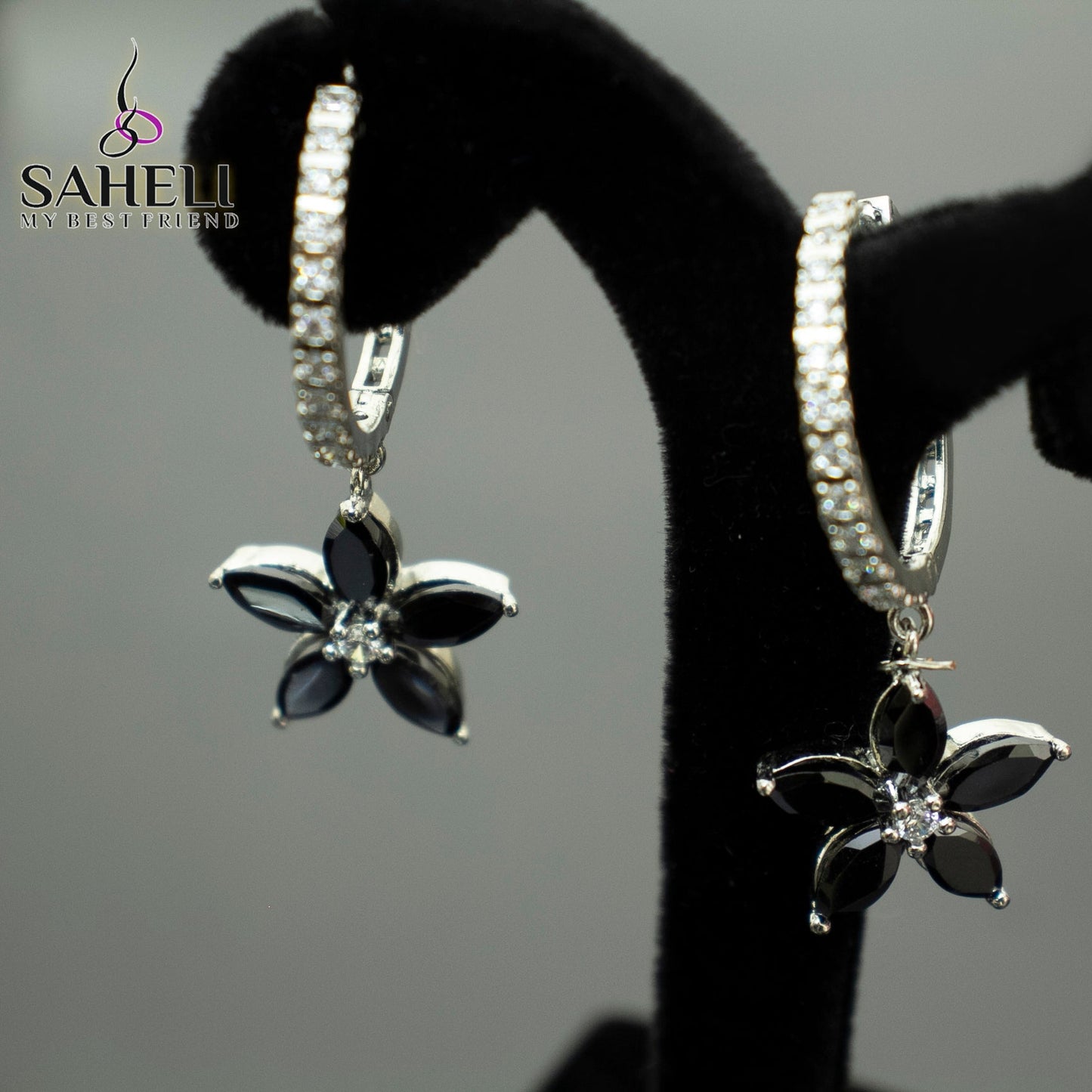 Black flower-shaped earrings with silver accents on a black stand against a gray background, branded 'Saheli'.