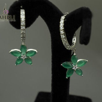 Flower-shaped earrings with green gemstones on a black stand against a gray background