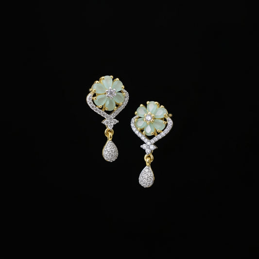 A pair of gold and white stone drop earrings with flower-shaped studs and teardrop charms, displayed on a dark gray velvet jewelry stand against a dark gray background.